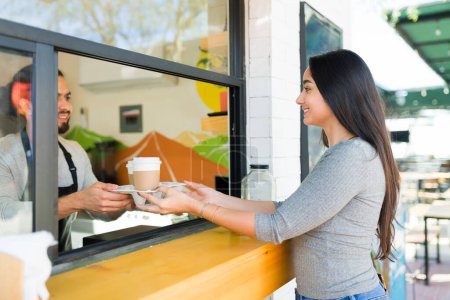 Photo for Beautiful young woman buying coffee to-go at the cafe and smiling at the barista looking happy - Royalty Free Image