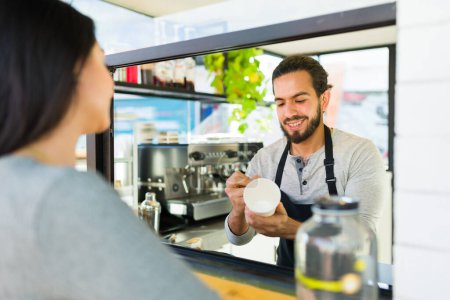 Téléchargez les photos : Heureux hispanique mâle barista souriant et parlant à un client tout en écrivant le nom sur le café à emporter - en image libre de droit