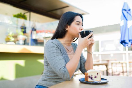 Téléchargez les photos : Attrayant hispanique jeune femme boire du café ou cappuccino et manger de la pâtisserie tout en se relaxant au café - en image libre de droit