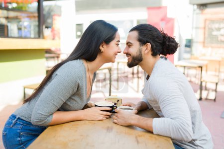 Foto de Perfil de una atractiva pareja hispana feliz a punto de besarse y sonreír mientras toma un café en la cafetería al aire libre - Imagen libre de derechos