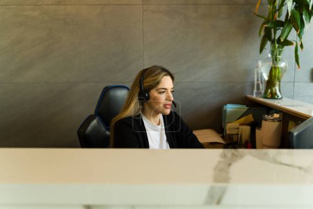 Téléchargez les photos : Belle réceptionniste caucasienne assise à la réception et portant un appareil mains libres - en image libre de droit