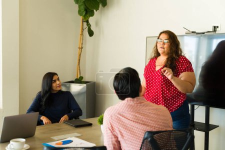 Foto de Mujer bastante latina con sobrepeso liderando una reunión de negocios y compartiendo algunas ideas con otros - Imagen libre de derechos