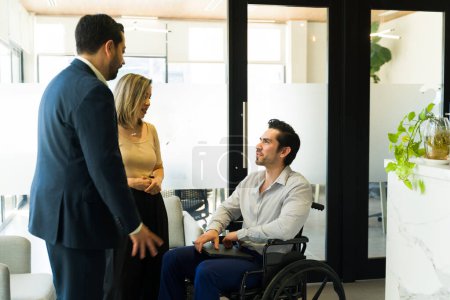 Téléchargez les photos : Groupe de gens d'affaires avec un handicapé en fauteuil roulant ayant une discussion amicale dans un couloir - en image libre de droit