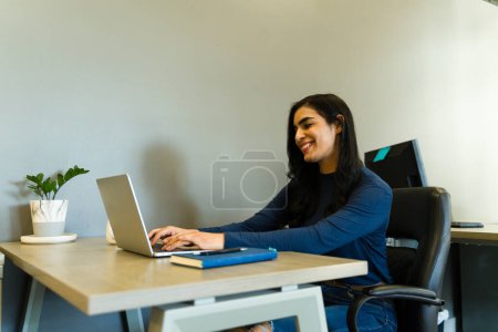 Photo for Beautiful Latin businesswoman sitting in an office and working on a laptop computer with a smile - Royalty Free Image