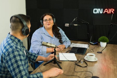 Téléchargez les photos : Belle hôte femme avec écouteurs sur son podcast parlant avec un invité lors de son émission de radio au studio insonorisé - en image libre de droit