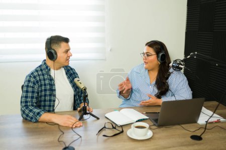 Photo for Hispanic woman and man working as radio co-hosts talking during a podcast show on the broadcasting station - Royalty Free Image