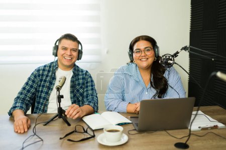 Photo for Attractive woman and man smiling while making a podcast and talking during a radio show on the microphone wearing headphones - Royalty Free Image