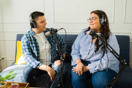 Photo for Hispanic man and woman co-hosts with headphones talking about a story while recording a podcast episode together - Royalty Free Image