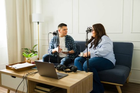 Photo for Latin woman and man drinking coffee while recording a talk show about fun stories during a podcast - Royalty Free Image