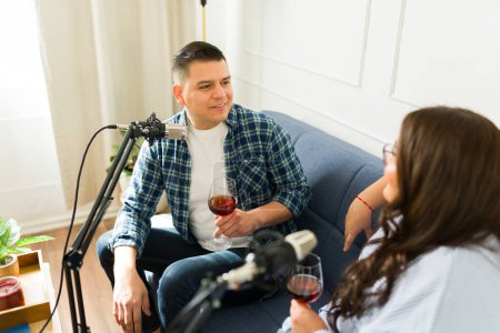 Foto de Latina podcast host sonriendo mientras habla con su invitado disfrutando de beber vino durante un talk show con un micrófono - Imagen libre de derechos