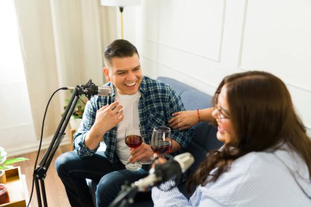 Photo for Attractive happy man laughing and having fun while talking with a microphone while recording a podcast with a woman guest drinking wine - Royalty Free Image
