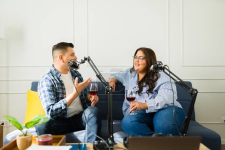 Foto de Mujer feliz y hombre emisoras beber vino y hablar de una historia divertida al micrófono mientras se graba un podcast - Imagen libre de derechos