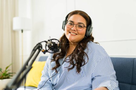 Foto de Hermosa mujer hispana sonriendo con auriculares mientras está feliz de grabar un episodio de podcast para su programa de entrevistas - Imagen libre de derechos