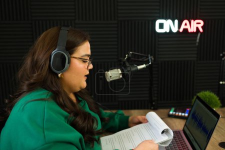 Photo for Latin woman with headphones recording podcast show in the radio studio while reading a script - Royalty Free Image