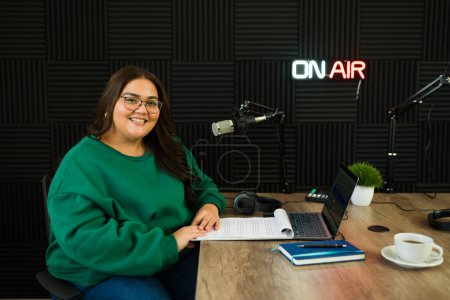 Téléchargez les photos : Belle femme hispanique hôte souriant faire un contact visuel tandis que dans le studio d'enregistrement insonorisé dans la station de radio - en image libre de droit