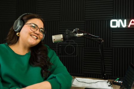 Photo for Hispanic woman host and broadcaster smiling with headphones and talking during a radio podcast episode - Royalty Free Image