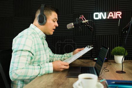 Photo for Hispanic man with headphones recording a podcast episode or hosting a radio talk show in the studio reading a script - Royalty Free Image