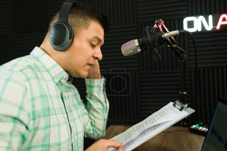 Photo for Latin attractive man on the radio during a talk show reading from a script while recording a podcast episode - Royalty Free Image