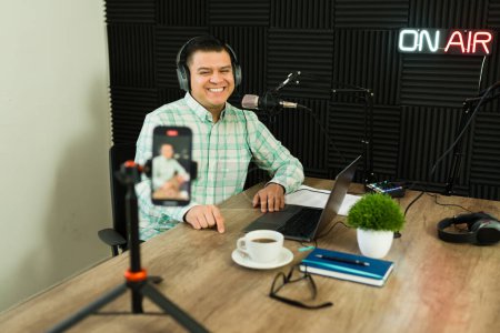 Téléchargez les photos : Heureux homme attrayant souriant en studio lors d'une vidéo en direct sur le smartphone tout en enregistrant un épisode de podcast - en image libre de droit