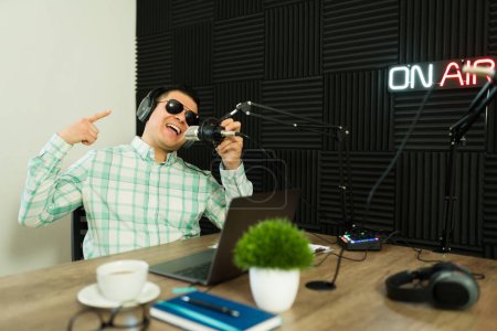 Photo for Funny man working as a broadcast or podcast host wearing sunglasses and recording an episode story in the studio - Royalty Free Image