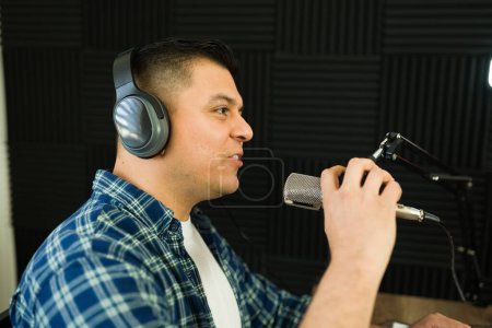 Téléchargez les photos : Profil d'un homme hispanique dans la trentaine avec des écouteurs parlant au microphone enregistrant un épisode de podcast ou une émission de radio - en image libre de droit