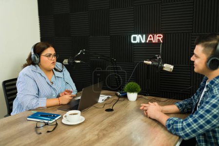 Téléchargez les photos : Femme latine dans le studio insonorisé parlant avec son invité lors d'une émission de radio ou lors de l'enregistrement d'un épisode de podcast - en image libre de droit