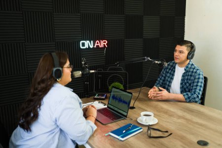 Photo for Hispanic woman broadcast on her talk show having an interview with a guest during her podcast at the recording studio - Royalty Free Image