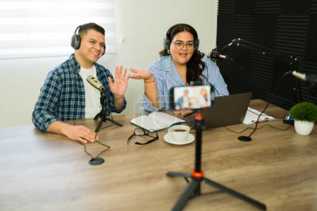 Téléchargez les photos : Heureux podcast hispanique co-hôtes souriant faire un flux en direct en ligne en utilisant le smartphone tout en enregistrant leur podcast - en image libre de droit