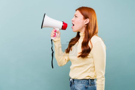 Téléchargez les photos : Profil d'une jolie femme rousse avec un mégaphone criant un message fort en colère dans un fond bleu - en image libre de droit