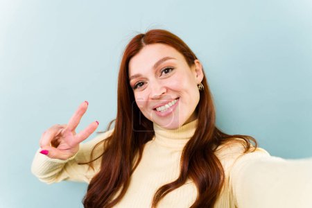 Téléchargez les photos : Perspective personnelle d'une femme séduisante aux cheveux roux faisant le signe de paix tout en prenant un selfie pour les médias sociaux - en image libre de droit