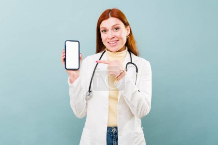 Photo for Cheerful attractive redhead doctor smiling pointing her finger to the smartphone while texting using social media - Royalty Free Image