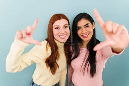 Photo for Gorgeous mexican and redhead women friends making a frame with their hands while taking a photo for social media - Royalty Free Image