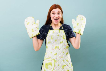 Caucasian redhead woman looking excited about cooking and baking wearing an apron and oven mitts