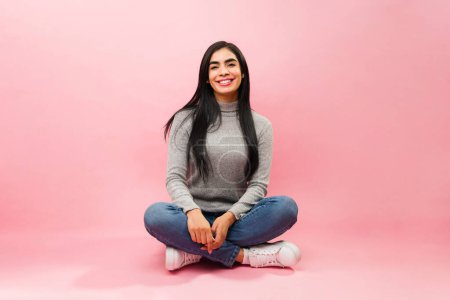 Photo for Relaxed hispanic woman in her 30s relaxing while sitting and smiling looking happy against a pink studio background - Royalty Free Image
