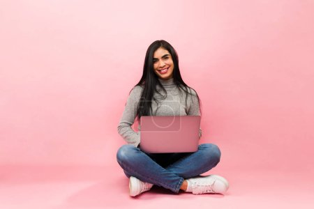 Photo for Cheerful mexican woman smiling sitting on the floor while working using her laptop and doing online shopping - Royalty Free Image