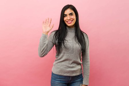 Photo for Beautiful hispanic woman in her 30s smiling and waving hello and saying hi in front of a pink background - Royalty Free Image