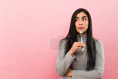 Photo for HIspanic woman using a fork and eating delicious food looking hungry and with cravings in a pink studio background - Royalty Free Image
