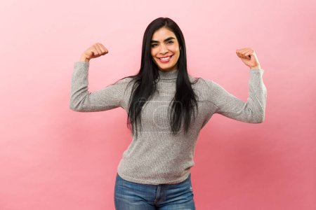 Foto de Mujer latina fuerte haciendo rizos bíceps y mostrando su fuerza promoviendo el poder de la chica en un fondo de estudio rosa - Imagen libre de derechos