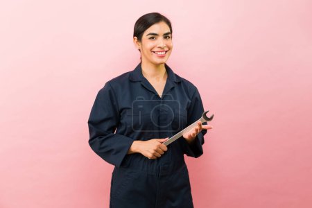 Foto de Hermosa mujer latina que trabaja como mecánico de coche con monos azules y el uso de herramientas mientras sonríe mirando feliz - Imagen libre de derechos