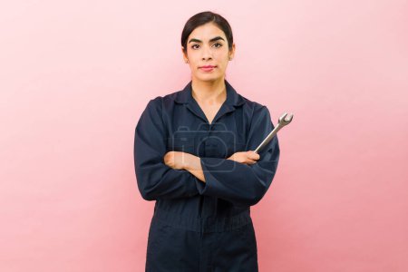 Foto de Mujer hispana con mono azul que usa herramientas mientras trabaja como mecánico de automóviles frente a un fondo rosa - Imagen libre de derechos