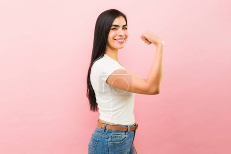Foto de Latina feliz joven sonriendo sintiéndose fuerte haciendo un rizo bíceps y promoviendo el poder de la chica en un fondo rosa - Imagen libre de derechos