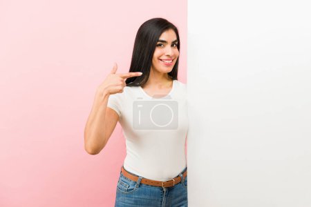 Téléchargez les photos : Magnifique hispanique jeune femme souriant avec des vêtements décontractés pointant son doigt vers un blanc espace de copie annonce - en image libre de droit