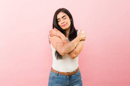 Foto de Amar a la atractiva mujer hispana sonriendo mientras se abraza promoviendo el amor propio o la autoestima frente a un fondo rosa - Imagen libre de derechos