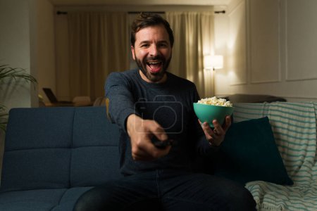 Photo for Excited man in his 30s smiling looking very happy while using the remote to watch a television show or movies while eating popcorn on the couch - Royalty Free Image
