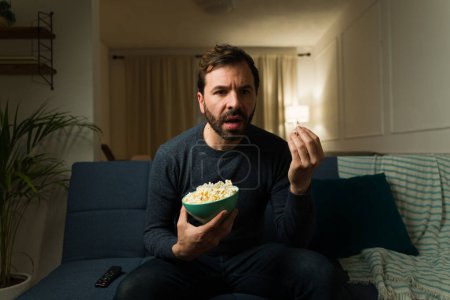 Foto de Hombre latino sorprendido disfrutando viendo una película de suspenso en la televisión mientras come palomitas de maíz en la sala de estar - Imagen libre de derechos