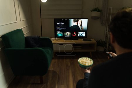 Foto de Caucásico hombre visto desde atrás sentado en el sofá en la sala de estar en busca de una película en la televisión utilizando un servicio de streaming - Imagen libre de derechos