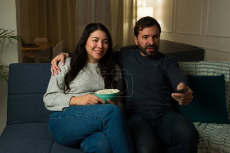 Photo for Beautiful happy couple eating popcorn while watching a tv show together while relaxing on the couch at night - Royalty Free Image
