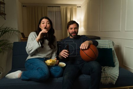 Téléchargez les photos : Marié heureux couple boire de la bière et profiter de regarder un match de basket-ball à la télévision dans le salon - en image libre de droit
