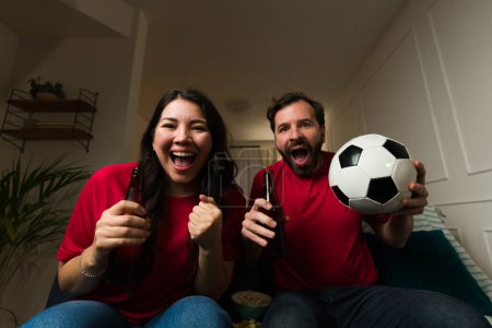 Téléchargez les photos : Excité attrayant couple marié avoir plaisir à regarder un match de basket à la télévision tout en célébrant leur équipe sportive préférée à la maison - en image libre de droit