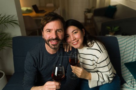 Photo for Attractive caucasian married couple enjoying drinking a glass of wine together during a relaxing date at home looking happy - Royalty Free Image